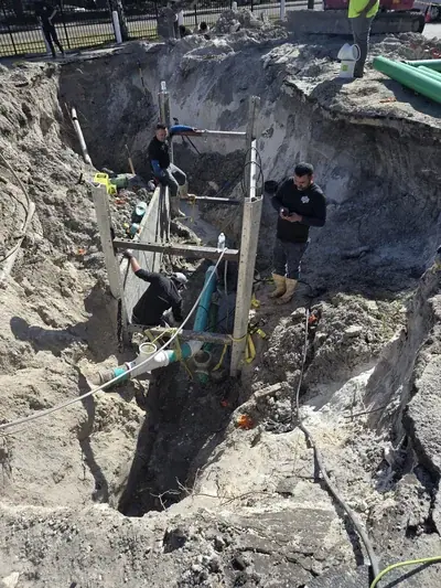 A large construction trench is dug into the ground, with several workers inside and around it. Green and white pipes are visible within the trench, supported by shoring.