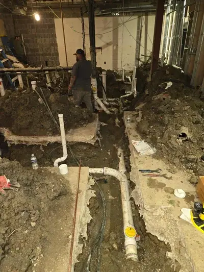 An indoor construction site with trenches dug into the dirt floor, revealing white PVC plumbing pipes. A worker in a dark shirt and cap stands in a trench, while another worker is visible on a ladder in the background.