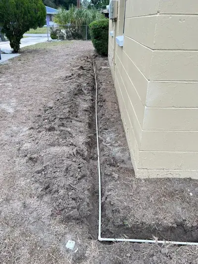 A narrow trench is dug in the dirt next to a light tan concrete block building, with a white pipe laid inside the trench and along the building's base. The surrounding ground is mostly bare dirt with some dry grass.