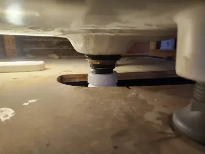 A low-angle view shows the underside of a white sink or toilet, with its drain assembly extending through a circular hole in a light brown wooden floor. The drain features brass-colored rings and connects to a white PVC pipe below the floor, with a gray pipe visible on the right.