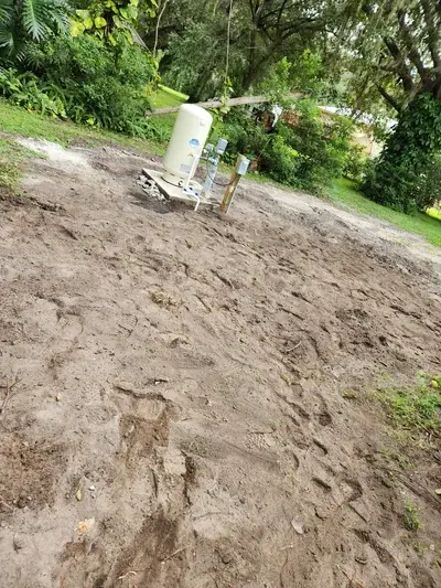 A light-colored water pressure tank stands on a concrete slab in a cleared dirt area. The ground is covered in footprints, with dense green vegetation and trees in the background.