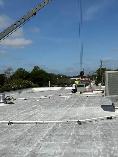 Two workers on a flat rooftop are installing an HVAC unit, with a crane lowering another unit into place. The roof is covered in light-colored material, and trees are visible in the background under a blue sky.
