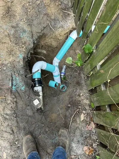 An overhead view shows a trench in the dirt with white and teal PVC plumbing pipes, some connected and some loose. A person's feet in brown work boots are visible at the bottom, next to a wooden fence on the right.
