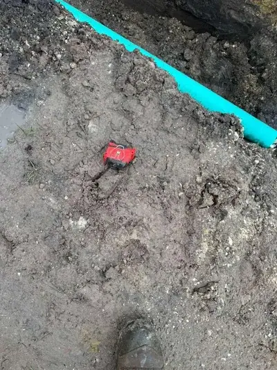 A muddy ground with a bright blue pipe running diagonally in the background. A small red and black object is partially buried in the mud, with a muddy boot visible in the bottom right.